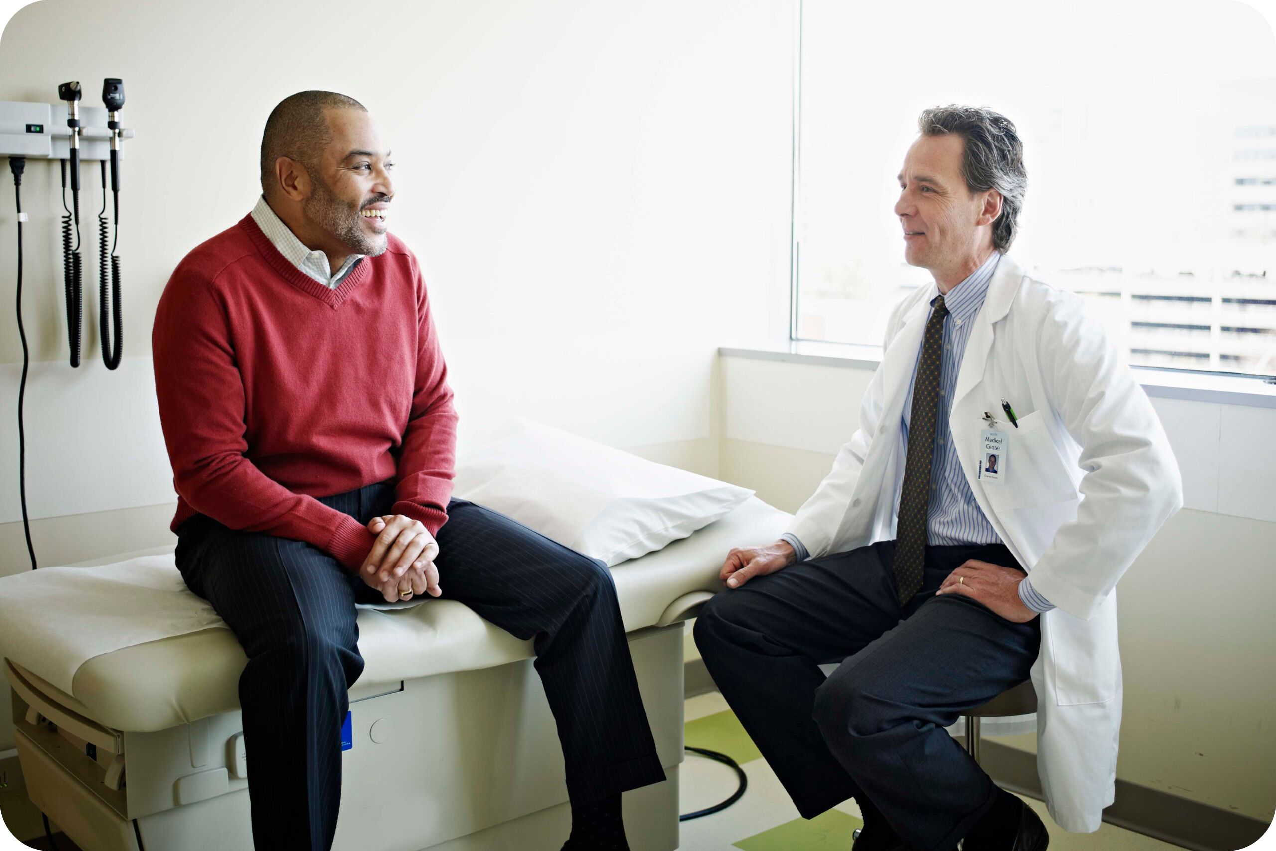 Older Black male patient sits on an exam table as he talks to a middle age White male doctor.