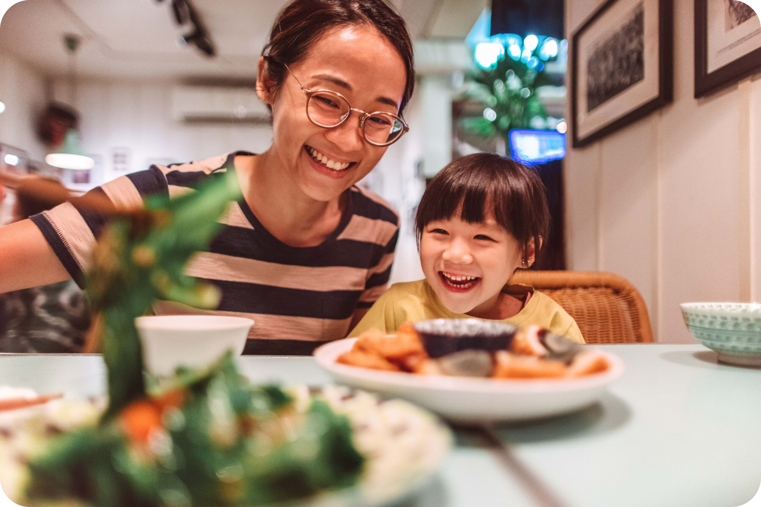 Young Asian mom serving vegetables to her smiling daughter while having meal.